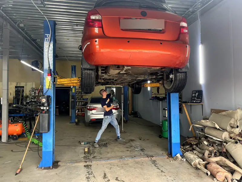 Оранжево-красная Skoda Fabia поднята на подъёмнике в боксе «Выхлопухи», мастер работает с выхлопом снизу