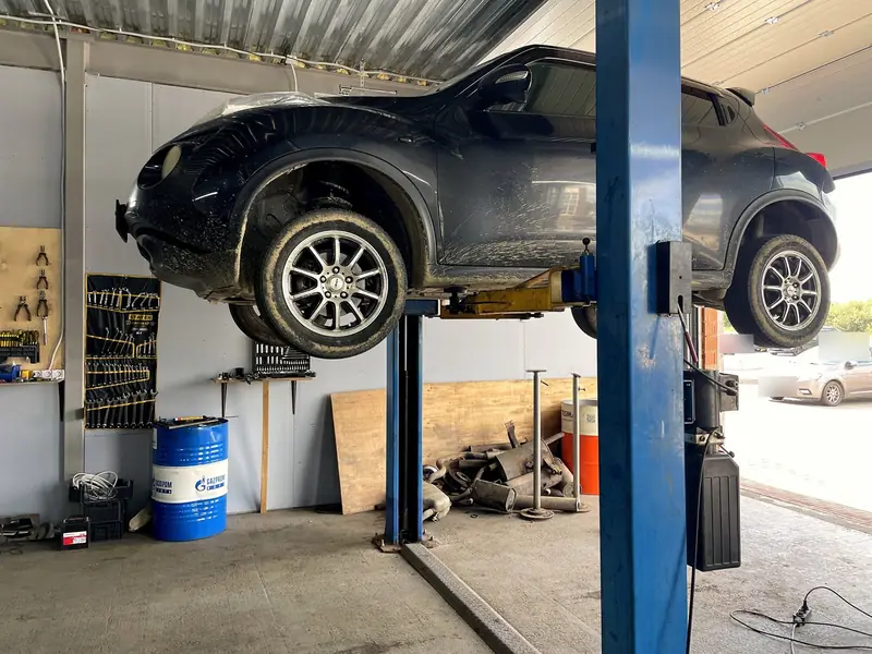 Чёрный Nissan Juke поднят на двухстоечном подъёмнике в боксе «Выхлопухи», вид сбоку, под ним на полу лежат снятые узлы выхлопа