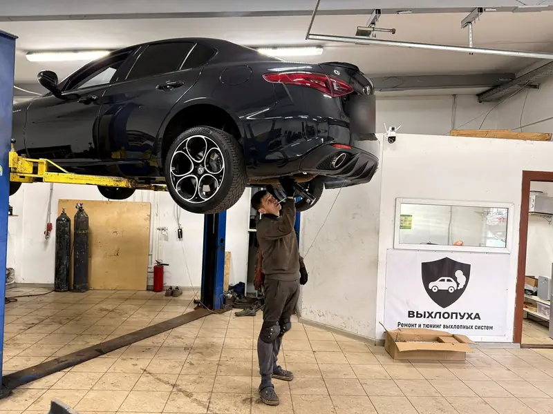 Чёрная Alfa Romeo Giulia на подъёмнике в цеху «Выхлопухи», мастер под задней частью, на стене баннер «Выхлопуха»