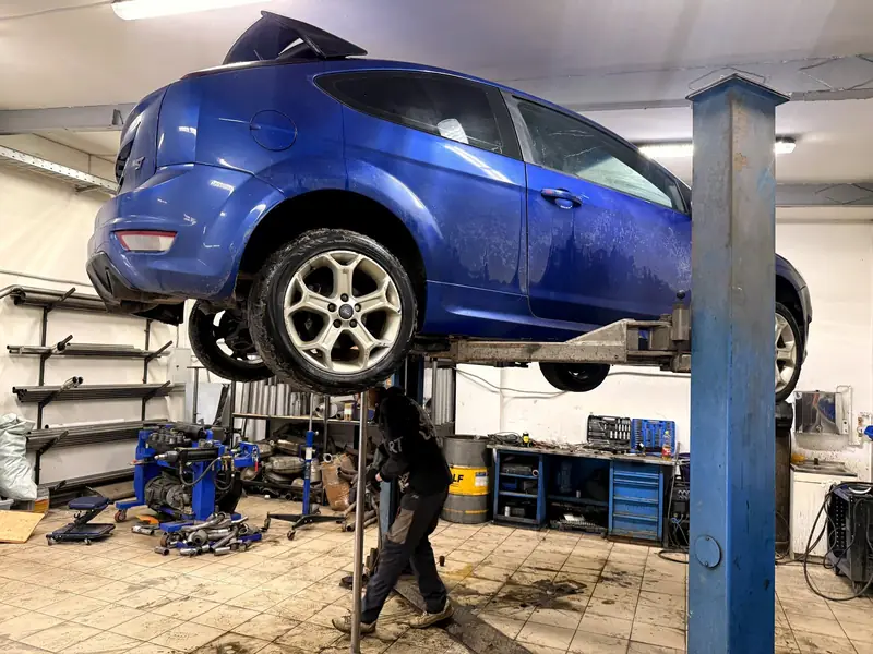 Синий трёхдверный Ford Focus ST на подъёмнике в цеху «Выхлопухи», мастер работает под машиной