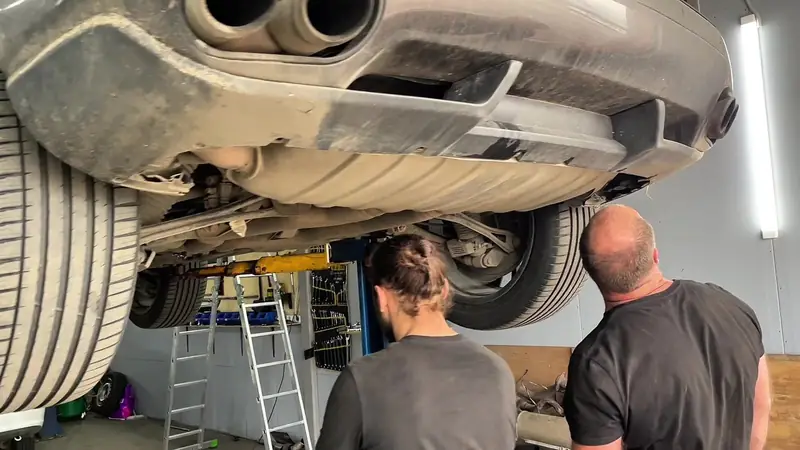 Porsche Cayenne Turbo на подъёмнике в цеху «Выхлопухи», мастера смотрят снизу, рядом снятые узлы старого выхлопа
